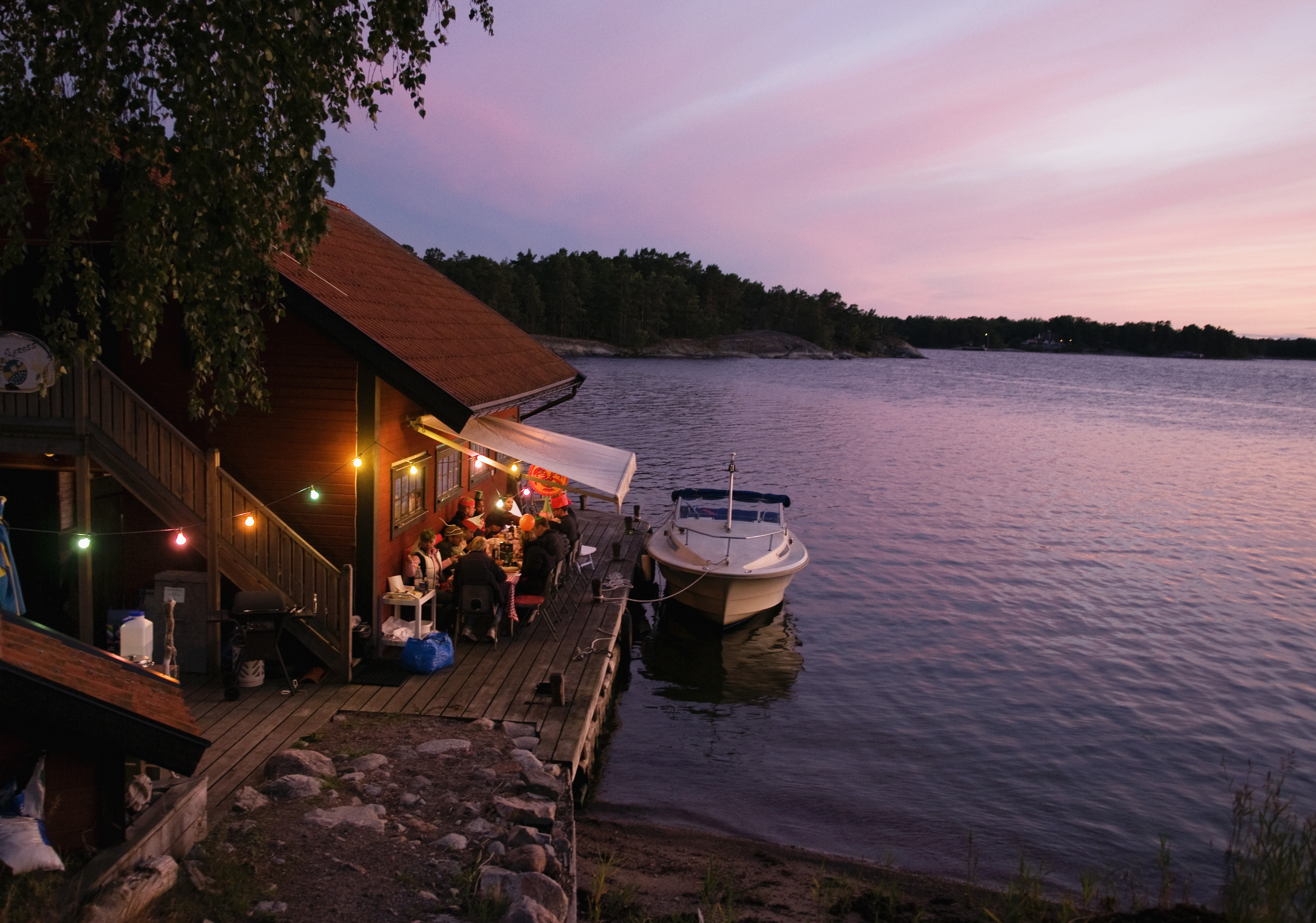 Flera personer sitter vid ett bord på en brygga. I vattnet intill bryggan ligger en båt.